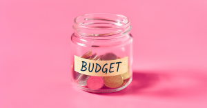 A glass jar labeled "BUDGET" filled with coins, placed against a pink background.