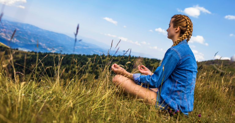 Woman meditating in a grassy field with mountains in the background.