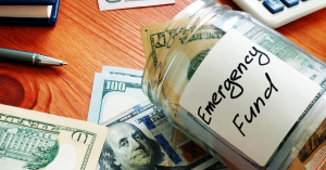 Jar labeled "Emergency Fund" filled with cash and surrounded by money on a wooden table.