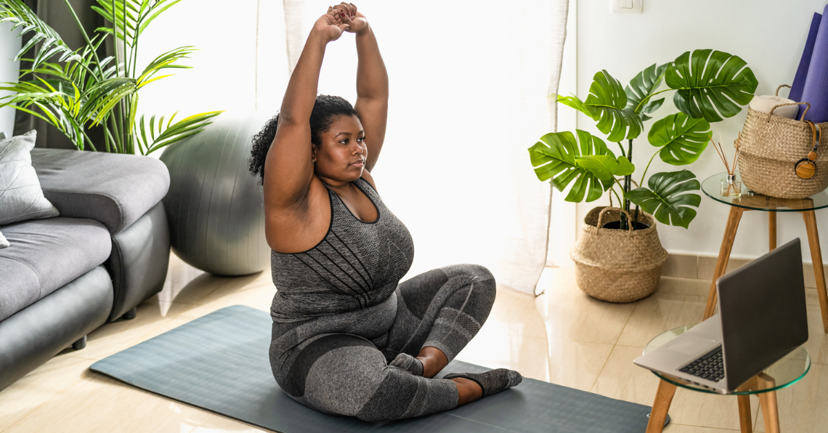 Woman practicing yoga at home on a mat, in a living room with plants and a laptop.