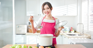 Woman cooking in a bright kitchen, smiling and holding a ladle and pot lid.