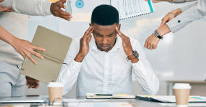 Man overwhelmed with work stress, holding his head while surrounded by multiple tasks and documents.