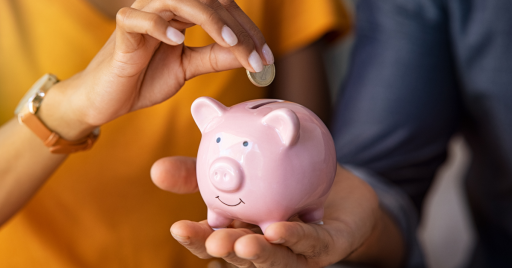 Close-up of hands placing a coin into a pink piggy bank, symbolizing saving money.